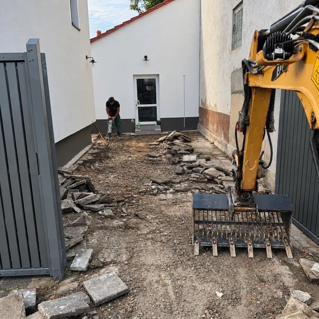 Minibagger mit Siebschaufel und Handwerker mit Abbruchhammer entfernen alte Platten in einem schmalen Hof