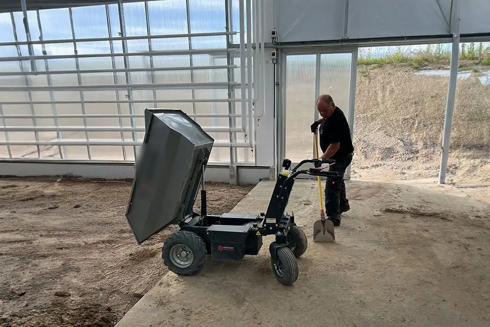 Handwerker mit Schaufel arbeitet mit einem Elektro-Dumper beim Materialtransport im Gewächshaus