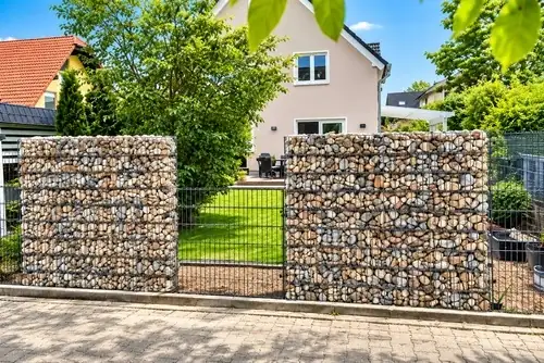 Zwei Gabionenelemente mit bunter Steinfüllung kombiniert mit einem anthrazitfarbenen Doppelstabmattenzaun vor einem Garten