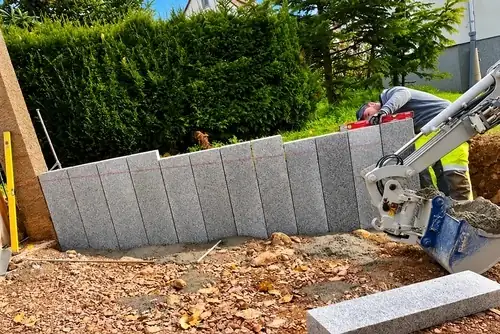 Granit-Palisaden werden als Hangkante in Beton gesetzt und per Wasserwaage ausgerichtet, rechts bringt ein Minibagger Beton