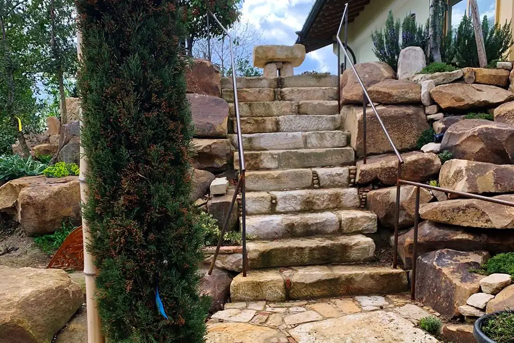 Treppe aus massiven Sandstein-Blockstufen zwischen Findlingsmauern, mit schlankem Eisengeländer und Natursteinbelag am Fuß