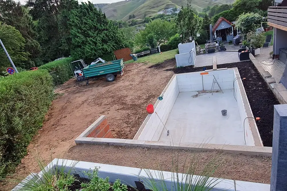 Rohbau eines Pools aus Schalsteinen auf Betonbodenplatte im Garten, daneben planierte Erdfläche, Minibagger und Anhänger