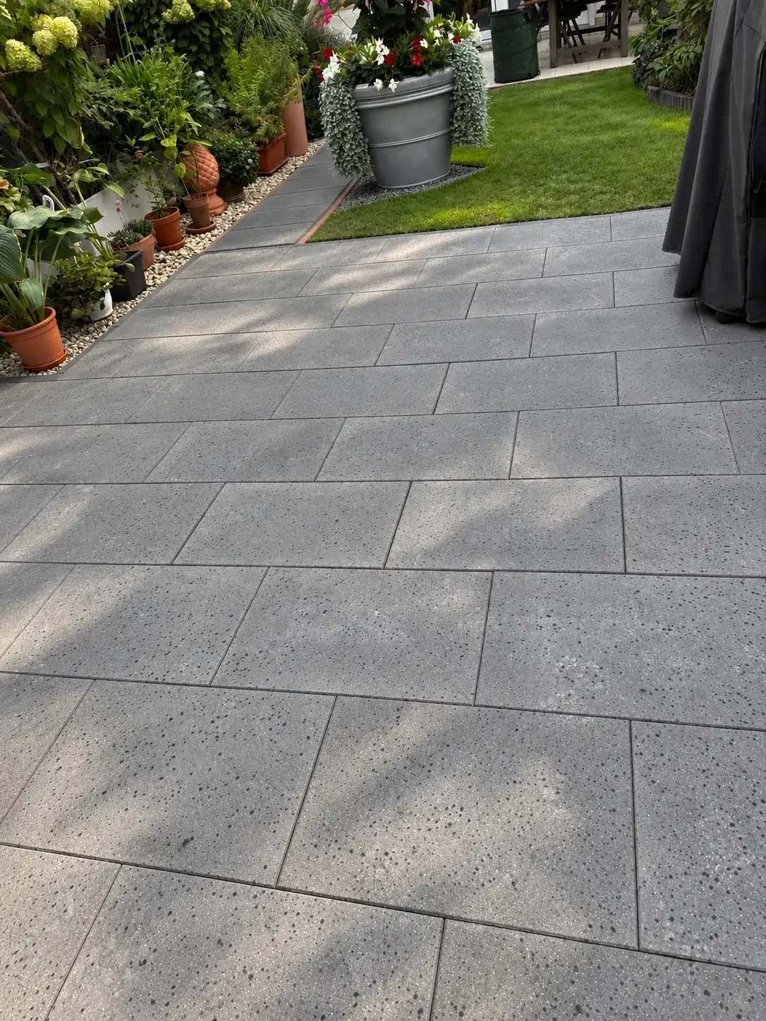 Fertige Terrasse aus großformatigen grau gesprenkelten Betonplatten, daneben Rasen, Kiesstreifen und Kübelpflanzen